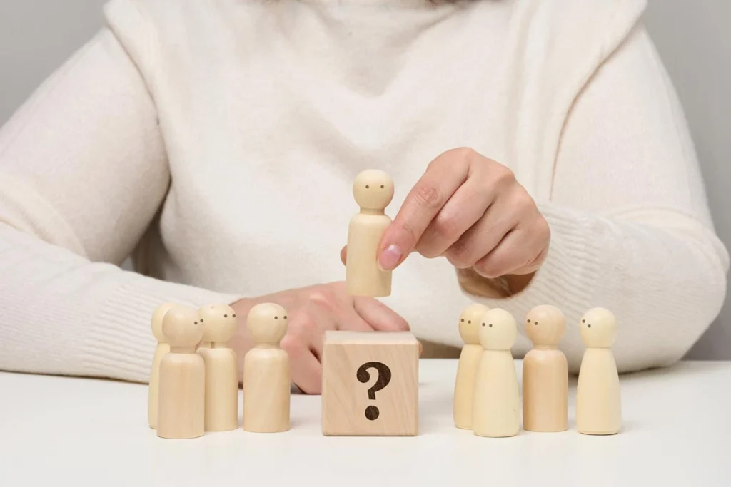 A woman’s hand lifting a wooden figure above other wooden figures and a question mark block, symbolizing Family Constellation therapy and emotional healing.