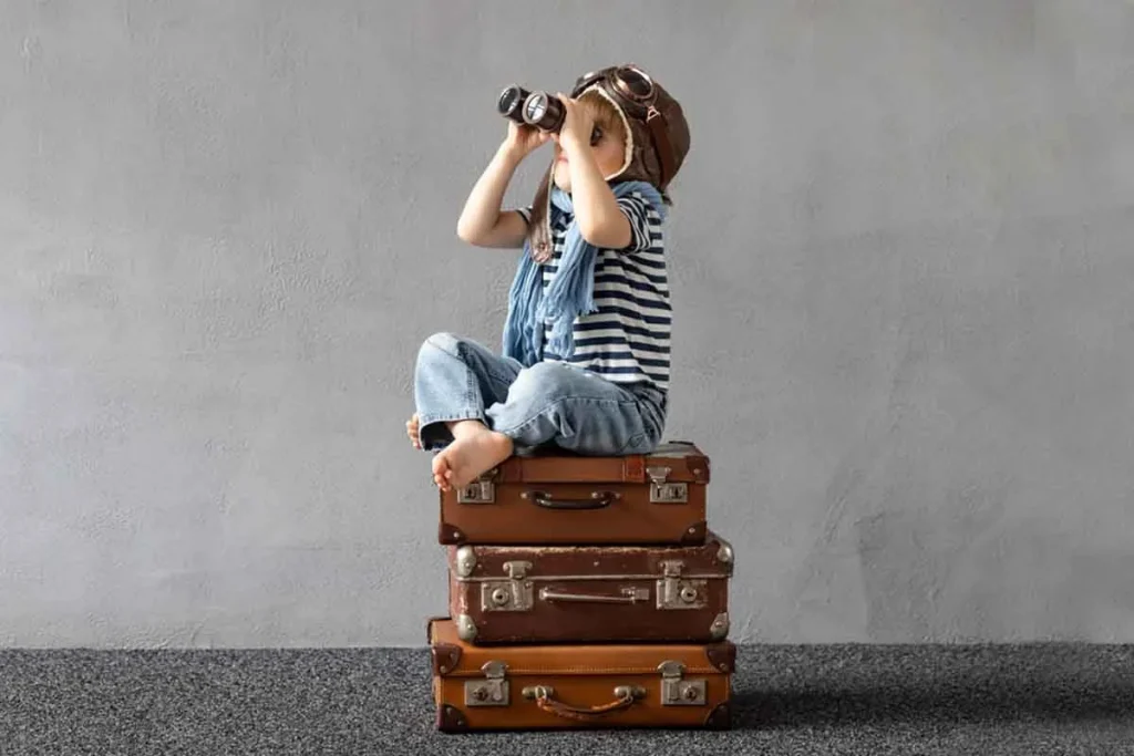 Little boy sitting on vintage suitcases looking at the sky with binoculars, symbolizing family travel, adventure, and awareness.