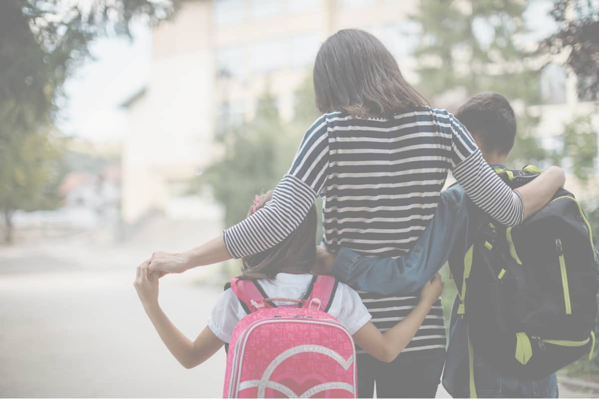 Mother walking with her two children after school, arms around both kids wearing backpacks